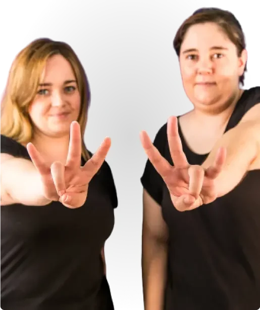 Dues noies miren a càmera, duen posada una samarreta negra, el fons és blau. Una aixeca el braç dret i l’altra l’esquerre i fan el signe BINOMILS.